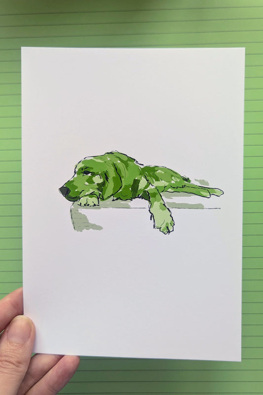 A hand holds a white art print featuring a green watercolor-style illustration of a dog lying down, set against a green lined notebook background.