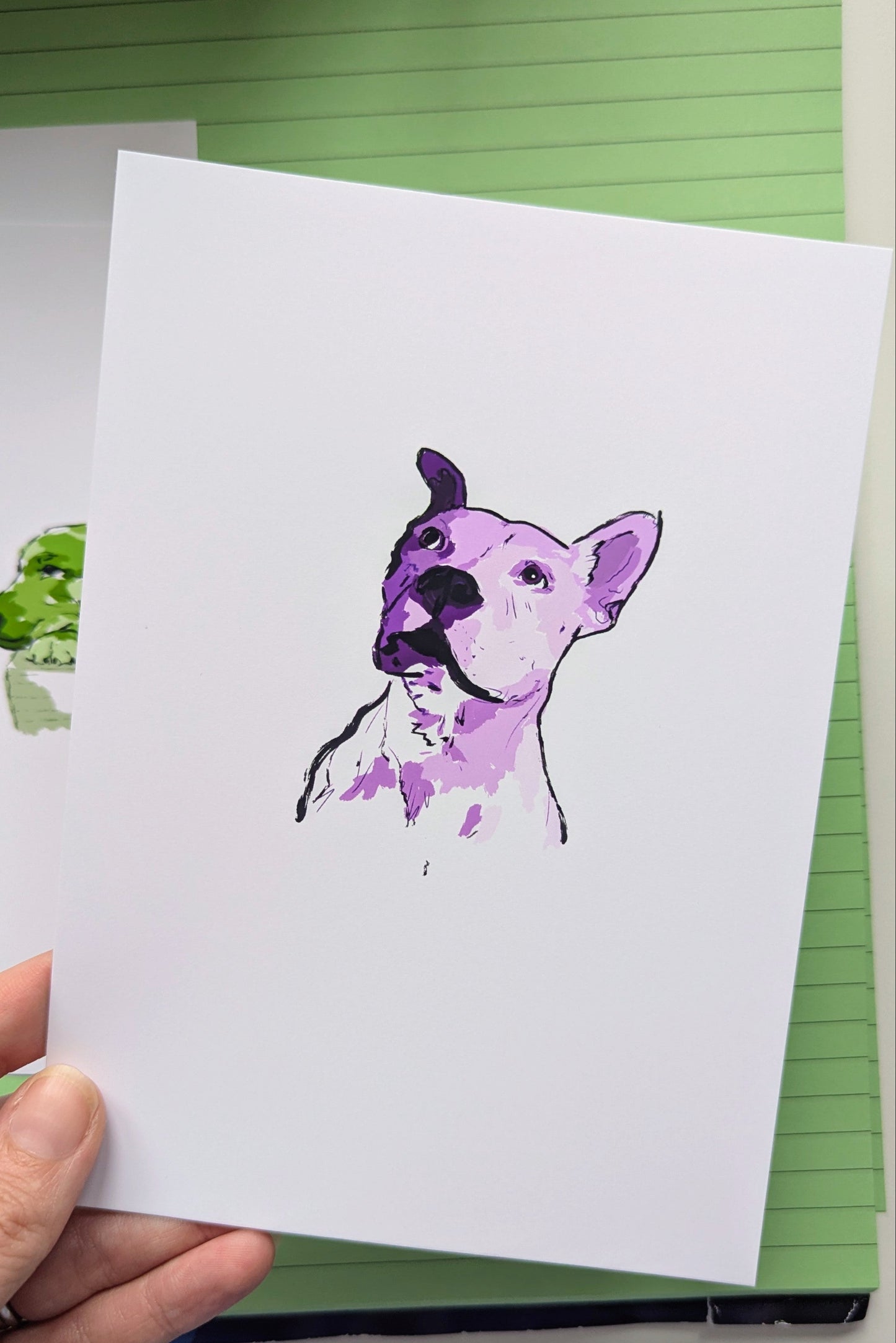 A hand holding a white art print featuring a watercolor-style purple and black illustration of a dog's head, set against a green lined notebook background.